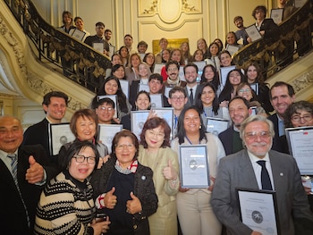 Grupo numeroso de personas sonrientes, adultos y jóvenes, posando en una escalera ornamentada, la mayoría sostiene certificados enmarcados