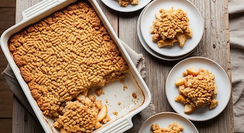 Vista cenital de un crumble de pera y manzana en fuente rectangular blanca, con algunas porciones en platos pequeños sobre mesa de madera rústica.