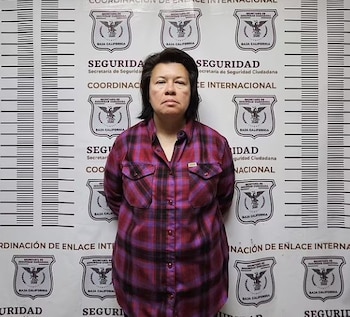 Una mujer con camisa a cuadros permanece de pie frente a un muro con logotipos de la Secretaría de Seguridad Ciudadana de Baja California
