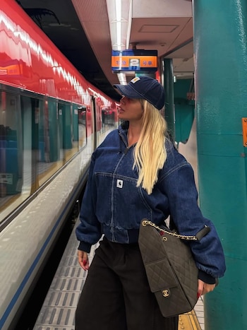 Wanda Nara de perfil en una plataforma de metro, con cabello rubio largo, gorra azul, chaqueta vaquera y un bolso acolchado, mirando un tren rojo
