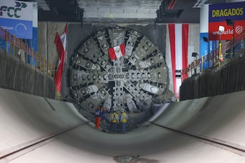 Vista frontal de la tuneladora "Delia" con la bandera de Perú, emergiendo de un túnel. Cuatro trabajadores están de pie frente a ella, bajo banderas rojas y blancas