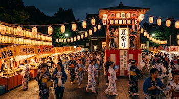 Personas con trajes tradicionales caminan entre puestos de comida iluminados por faroles de papel junto a una torre de madera de noche.