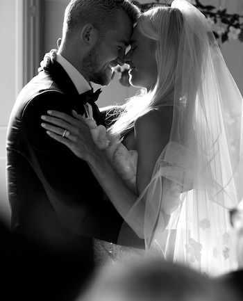 Fotografía en blanco y negro de un hombre con esmoquin y una mujer con velo y vestido de novia que se abrazan con los rostros juntos