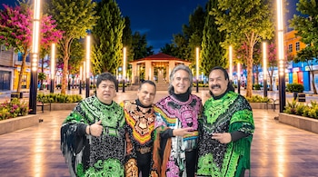 Cuatro hombres vestidos con ponchos tradicionales bordados posan en un paseo arbolado con farolas encendidas al anochecer.