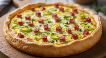 Tarta salada con base dorada, relleno cremoso con rodajas de puerro verde, cubos de jamón y hierbas aromáticas sobre una tabla de madera.