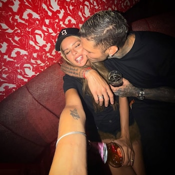 Hombre con tatuajes besa a mujer con gorra y piruleta. Ambos sostienen bebidas. Fondo rojo con figuras humanas. Brazo tatuado en primer plano