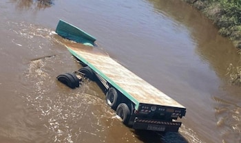 Vista aérea de un remolque de camión, de color verde y marrón, semisumergido en un arroyo de aguas turbias con vegetación en los márgenes