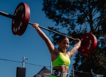 Una atleta vestida con indumentaria deportiva verde sostiene una barra con discos rojos levantada sobre su cabeza bajo un cielo despejado