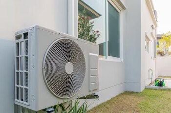 Unidad exterior de bomba de calor blanca montada en la pared de una casa moderna con ventanas, césped verde y una manguera en el fondo