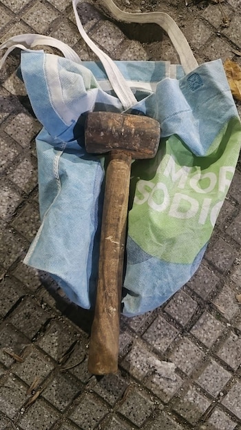 Un mazo de madera con cabeza y mango oscuros descansa sobre una bolsa de tela azul y verde con letras blancas, sobre un suelo de baldosas grises
