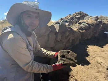 Un hombre con sombrero de ala ancha y guantes rojos sostiene una pieza de piedra perforada frente a un muro de pirca