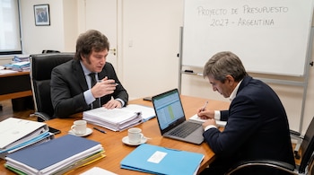 Javier Milei y Luis Caputo sentados ante una mesa con carpetas, tazas de café, una computadora portátil y una pizarra detrás.