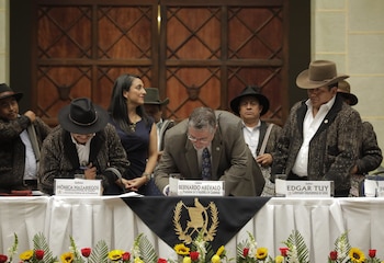 Mesa con personas, una persona firma documento, personas con sombreros, pancarta con escudo de Guatemala, flores rojas y amarillas, pared de madera, carteles de nombres