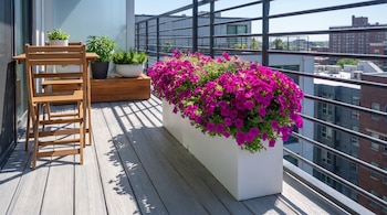Mesa y sillas plegables de madera junto a una gran jardinera blanca con flores magenta en un balcón con barandilla metálica.