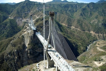 Vista aérea de un puente atirantado en construcción en un valle montañoso, con grúas, cables de acero, estructuras de concreto y vegetación