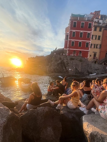 Grupo de personas sentadas en rocas frente al mar con un atardecer, dos botes, y edificios coloridos en un acantilado a la derecha