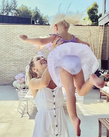 Una mujer levanta en el aire a una niña pequeña, ambas con ropa clara, en un patio exterior con un muro de ladrillos y globos decorativos