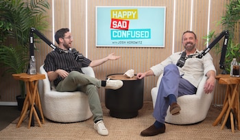 Josh Horowitz y Ben Affleck conversan sentados en sillones blancos frente a micrófonos dentro de un estudio