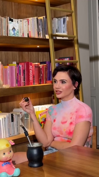 Celeste Cid sentada junto a una mesa con un mate, frente a una biblioteca con estantes llenos de libros y una escalera dorada