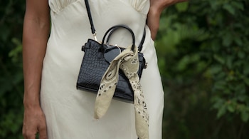 Primer plano de una persona llevando un bolso negro con textura de cocodrilo, un pañuelo bandana beige atado y gafas de sol, sobre un vestido crema.