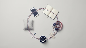 Vista cenital de objetos cotidianos en círculo sobre superficie gris claro: móvil, cuaderno, auriculares, café, pastillas y almohada, unidos por hilos azules y rojos.