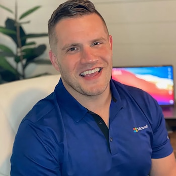 Hombre sonriendo a cámara con camisa polo azul y logo de Microsoft. Detrás se aprecian una planta de interior y un monitor de computadora