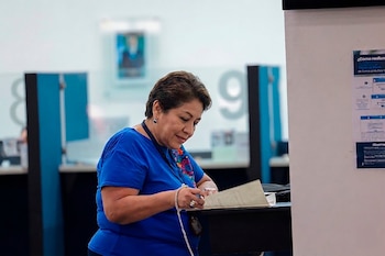 Una mujer de mediana edad con cabello oscuro, blusa azul y un collar colorido, revisa documentos con un bolígrafo en una oficina
