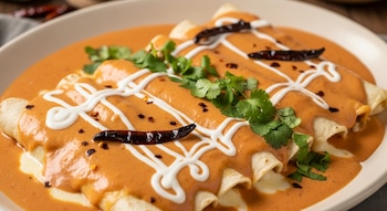 Primer plano de enchiladas cubiertas con una salsa naranja cremosa, decoradas con líneas de crema blanca, hojas de cilantro y chiles chipotle secos.