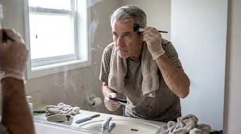 Primer plano de un hombre de mediana edad con guantes, aplicando tinte oscuro en su cabello gris con un cepillo frente a un espejo en el baño.