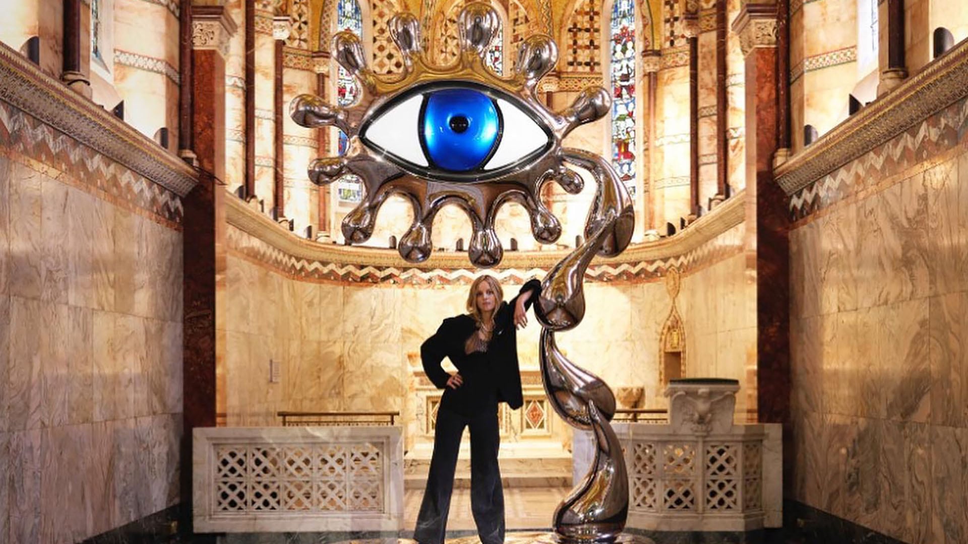 La artista Charlotte Colbert posa junto a su impactante escultura cromada en forma de ojo, exhibida dentro de un edificio con detalles arquitectónicos clásicos (@colbertcharlotte)