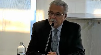 Un hombre con cabello gris, bigote y gafas, vestido con traje y corbata, habla ante un micrófono. Una botella de agua está visible en la mesa