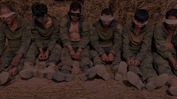 Seis hombres vestidos con ropa militar verde, sentados en el suelo, con vendas en los ojos y las manos atadas frente a un montón de paja