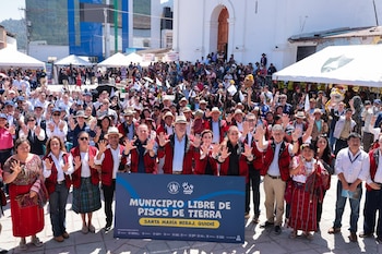 Gran grupo de personas sonrientes en un evento al aire libre, muchos con trajes tradicionales, alzando sus manos frente a un edificio blanco y un cartel azul