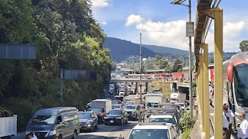 Vista panorámica de una carretera con denso tráfico vehicular y camiones parados; montañas y vegetación densa flanquean la vía bajo un cielo azul con nubes
