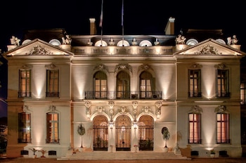 Fachada de un palacio de dos plantas iluminado de noche, con tres arcos de rejas en el ingreso y dos mástiles con banderas en el techo