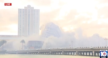 Destruição de um edifício desmoronando em uma nuvem de poeira. Prédios brancos e uma ponte sobre a água com palmeiras podem ser vistos