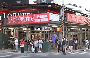 La fachada del restaurante Red Lobster en Times Square, Nueva York, está cubierta con andamios y banners rojos que anuncian su apertura durante la construcción. Varias personas transitan por la acera