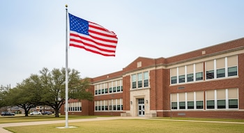 Vista exterior de una escuela de ladrillo rojo de dos pisos con ventanas, un asta con la bandera de Estados Unidos ondeando y un césped verde.