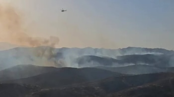 Un helicóptero oscuro vuela en un cielo claro sobre varias cadenas montañosas cubiertas de humo gris y marrón por un incendio