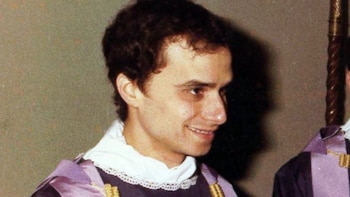 Primer plano de un hombre joven de cabello oscuro y sonriente que viste un hábito blanco y un ornamento litúrgico morado