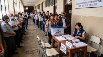 Una fila de personas en el pasillo de un colegio frente a una mesa con papelería electoral, una ánfora transparente y una trabajadora de la ONPE.