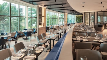 Interior de un restaurante moderno con grandes ventanales, mesas puestas, sillas azules y grises, y un muro verde, mostrando un ambiente amplio y luminoso
