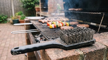 Un cepillo de cerdas de acero con mango negro sobre ladrillos junto a una parrilla con alimentos en cocción.