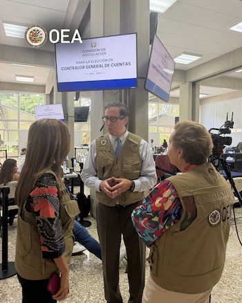 Tres personas con chalecos con el logotipo de la OEA conversan ante una pantalla sobre la elección de Contralor General de Cuentas