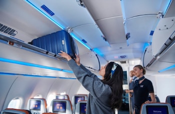 Cabina de avión con asientos, pantallas, compartimentos superiores, una mujer colocando una maleta azul y una auxiliar de vuelo