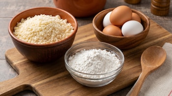 Imagen cenital de un tazón con queso sardo rallado, otro con almidón de mandioca y un tercero con huevos sobre una tabla de madera.
