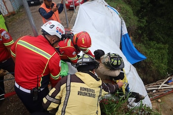 Cuatro bomberos con cascos y chaquetas amarillas y rojas asisten a una persona cerca de una lona blanca en una pendiente