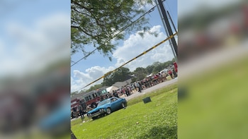 Un coche azul clásico, un camión de bomberos y cinta amarilla de precaución en una calle con una multitud de personas y césped verde