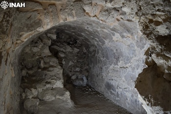 Pasillo interior de una estructura de piedra con techo abovedado, paredes de roca y suelo de tierra. Se observa el logo de INAH