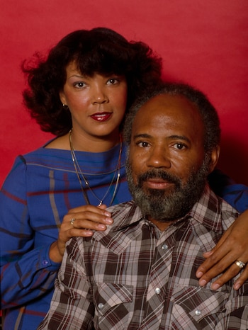 James Meredith vistiendo una camisa a cuadros sentado junto a una mujer con blusa azul sobre un fondo rojo